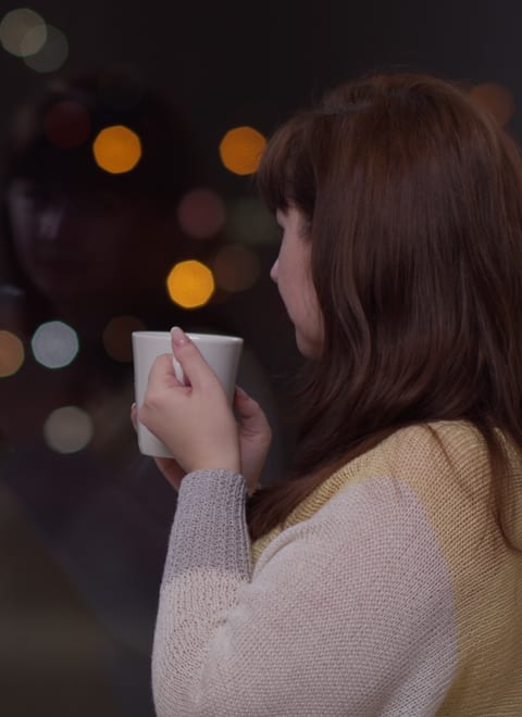 A parent pausing at a window with a hot drink